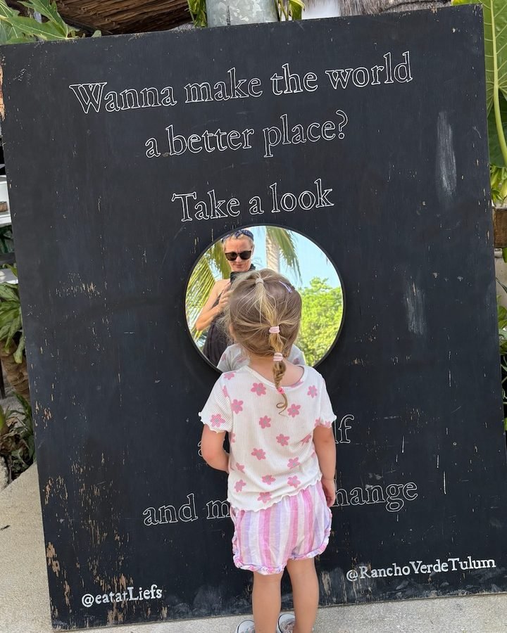 Young girl looking into the round mirror on the black Liefs chalkboard sign saying Wanna make the world a better place in Tulum